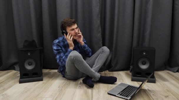 Muž ve sluchátkách poslouchá hudbu a zpívá. Mladý hudebník zpěv píseň ve sluchátkách pomocí notebooku, zatímco sedí na podlaze v domácím studiu — Stock video