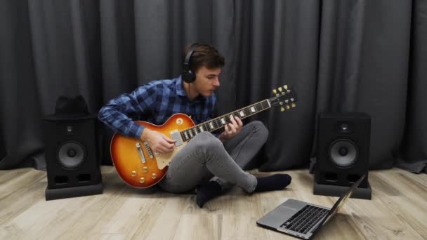 ギターを学ぶ男。ノートを使ってエレキギターを弾くヘッドフォンの若い男性。十代の若者たち座っています床にリビングルームと学習コードでギター — ストック動画