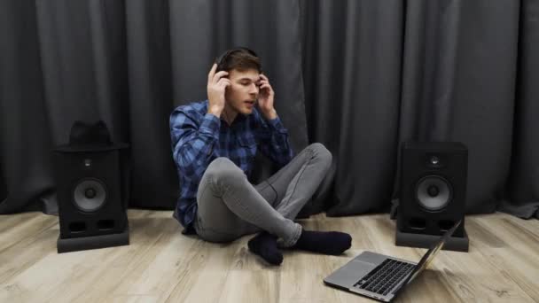 Mladý muž poslouchá hudbu ve sluchátkách a zpívá. Muž si nasadí sluchátka a začne zpívat, zatímco sedí na podlaze v moderním obývacím pokoji. Koncept životního stylu — Stock video