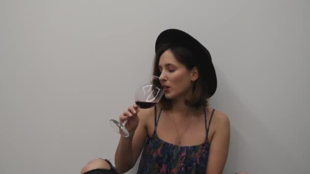 Yerde oturan ve kırmızı şarap içen bir kadın. Beyaz arka planda şarap içen bir kadın. Parlak makyajlı şık bir kız şarap tadıyor ve gülümsüyor. — Stok video
