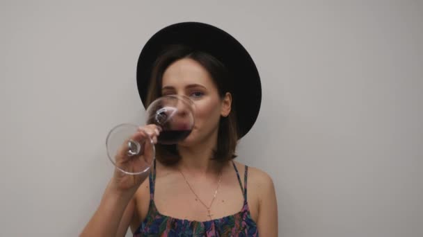 Сомелье пьет вино. Женщина пробует красное вино на белой стене. Стильная молодая девушка с ярким макияжем и шляпными напитками и глотает вино в студии — стоковое видео