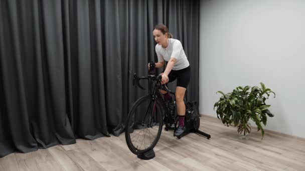 Woman on indoor smart trainer cycling drinks water or isotonic drink while riding a bicycle. Professional cycling training program — Stock Video