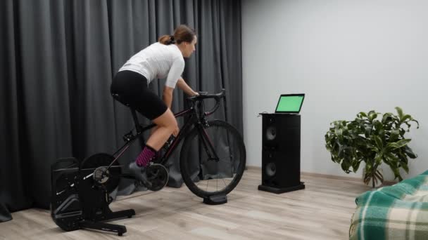 Mujer ciclista bebe agua mientras se entrena en el entrenador de bicicletas inteligentes en casa mirando la pantalla de la computadora. Concepto de ciclismo interior — Vídeo de stock