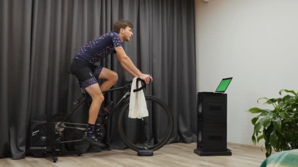 Der junge sportliche Kerl wischt mit dem Handtuch, während er auf dem stationären Smart-Fahrradtrainer radelt. Motivierte Mann hartes Training auf dem Fahrrad zu Hause. Konzept zum Abnehmen — Stockvideo