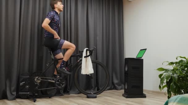 Mężczyzna zamykający koszulkę rowerową podczas treningu na inteligentnym rowerze treningowym wewnątrz. Młody mężczyzna intensywnie pedałuje i wykonuje ćwiczenia cardio i siłowe w domu stacjonarny trenażer rowerowy — Wideo stockowe