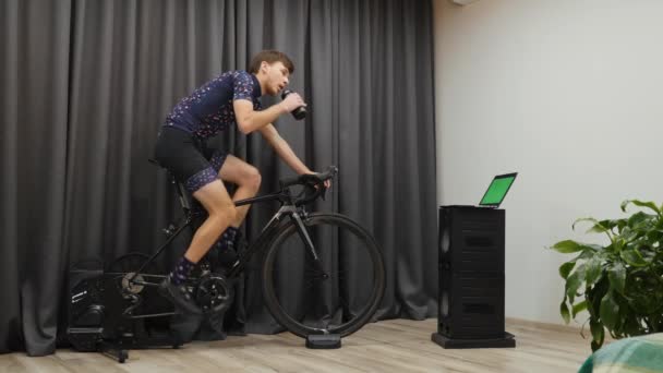 Junger sportlicher Mann trinkt beim Fahrradfahren auf einem Smart-Fahrradtrainer Energie. Motivierte Triathleten bereiten sich auf Triathlonwettkämpfe in der Halle vor. Mann tritt auf Fahrrad, um Gewicht zu verlieren — Stockvideo