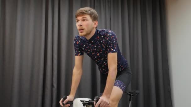 Facet jeździ na rowerze z siodła i pije izotoniczne podczas ciężkiego treningu na inteligentnym rowerze treningowym. Koncepcja wirtualnej jazdy na rowerze. mężczyzna robi ćwiczenia w domu — Wideo stockowe