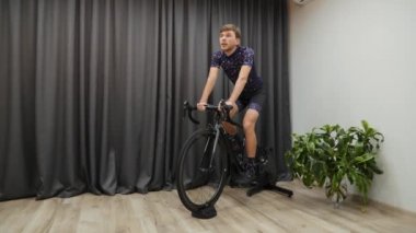 Bisiklet eğitmeniyle evde bisiklet sürmeye uygun bir erkek. Bisikletle içeride sıkı antrenman yapan bir sporcu. Sanal bisiklet konsepti. Kendine güvenen genç adam bisiklet sürerken akıllı antrenör