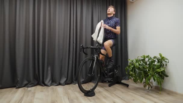 Professional cyclist training on bicycle trainer indoors. Male athlete hard pedaling on bike at home, sweating, holding towel and wiping. Man cycling virtual concept — Stock Video