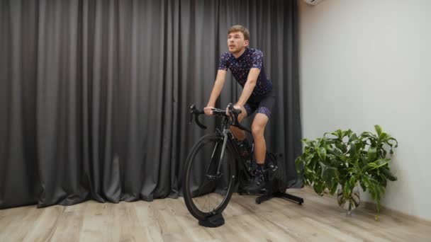 Fit male cycling at home on bicycle trainer. Athlete hard training on bike indoors. Virtual cycling concept. Young confident man pedaling on bike smart trainer — Stock Video