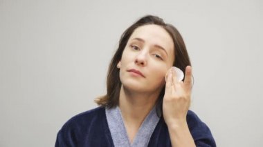 Kadın yüzünden makyaj yapıyor. Mavi bornozlu kadın kozmetik ürünleri yüzünden çıkarıp aynaya bakıyor. Makyaj kaldırılıyor. Sağlık ve güzellik kavramı.