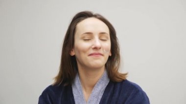 Gülümseyen genç bir kadın yüz kremi sürüyor. Mavi bornozlu kadın yüzüne gece kremi sürüyor ve uyumaya hazırlanıyor. Tatlı, çekici bir bayan yüzüne bakıyor ve anti-yaş kremi sürüyor.