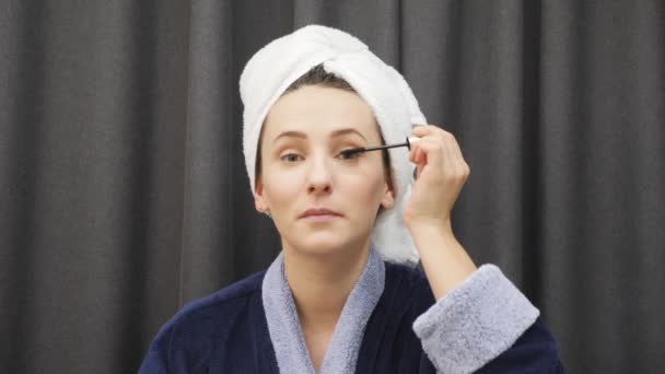 Jovem fêmea aplicando rímel em pestanas. Mulher de roupão azul e toalha na cabeça segurando rímel e fazendo maquiagem em casa. Mulher olhando para o espelho e colocando cosméticos no rosto — Vídeo de Stock
