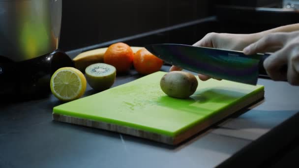 Mujer corta kiwi en la cocina con frutas frescas en el fondo. Kiwi verde de corte femenino por cuchillo sobre tabla de madera. Mujer preparando jugo fresco. Dieta saludable Alimento ecológico — Vídeo de stock