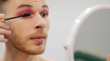 Kendine makyaj yapan travesti, kapat. Genç, gülümseyen çekici adam rimel sürüyor. Transseksüel aynaya bakıyor ve gülümsüyor