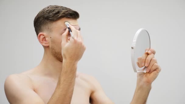 Chlápek, co se namaloval. Mladý pohledný muž dává na tvář maskér. Transsexuál nanáší make-up a dívá se do zrcadla. Travesty make-up koncept — Stock video