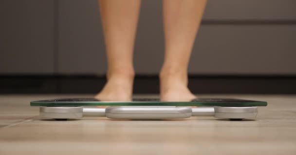 La mujer pisa básculas. Pies femeninos comprobando peso corporal en cocina. Piernas humanas midiendo peso, vista de cerca — Vídeo de stock