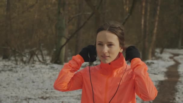 A mulher põe auscultadores. Mulher sorrindo bonito coloca fones de ouvido no parque antes de correr. Atleta profissional se preparando para o treinamento ao ar livre duro na floresta no inverno. Conceito de desporto — Vídeo de Stock