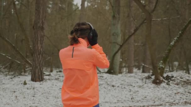 Frau beginnt im Park zu joggen. Frau in orangefarbener Jacke setzt Kopfhörer auf und beginnt im Winterwald zu laufen. Sportlerin ist hartes Training am Morgen als Teil des täglichen Trainings. Trailrun-Konzept — Stockvideo