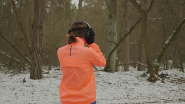 Mujer en auriculares comienza a correr en el parque. Mujer en chaqueta naranja con hermoso cuerpo corriendo en el bosque de invierno. Chica haciendo ejercicios de fitness al aire libre para bajar de peso. Entrenamiento intenso atleta — Vídeos de Stock