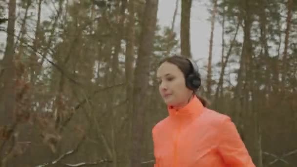 Kobieta biegająca w parku. Pewna siebie skupiła się na kobiecie w słuchawkach i pomarańczowej kurtce biegającej rano w zimowym lesie. Profesjonalny biegacz ciężko trenujący i przygotowujący się do półmaratonu — Wideo stockowe