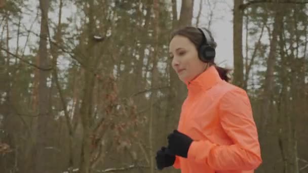 Kvinnlig jogging i vinterskogen. Motiverad koncentrerad kvinna i hörlurar som kör i parken. Professionell löpare idrottare gör utomhus träningspass i lågsäsong. Kvinna som tränar kondition. Kör koncept — Stockvideo