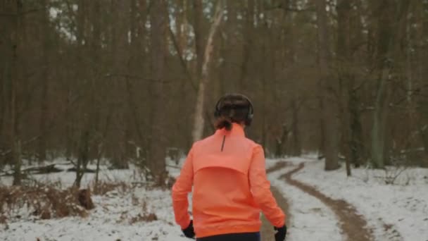 Femme dans les écouteurs a fini de courir des séances d'entraînement dans le parc. Face arrière suivre la vue de l'athlète féminine sportive en cours d'exécution dans le parc d'hiver. Fille arrête de courir et la respiration dure après des exercices de conditionnement physique intenses — Video