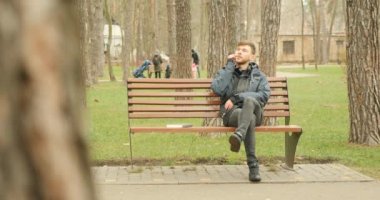 Genç sakallı adam arıyor ve telefonda konuşmaya başlıyor gülerek ve sohbetten zevk alarak sonbahar parkında bankta oturuyor..
