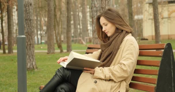 Kobieta czyta książkę siedzącą na ławce w jesiennym parku w brązowym okopie i szaliku. Koncepcja czytania — Wideo stockowe