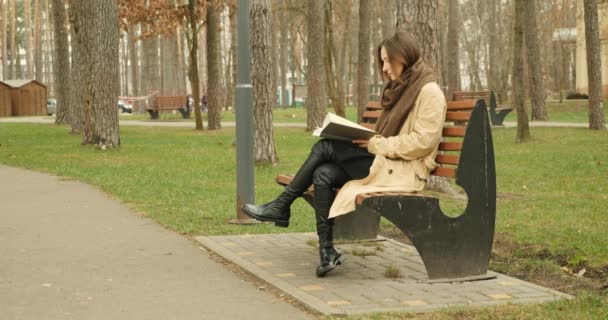 Donna che finisce di leggere un libro sulla panchina si alza in piedi e lascia il parco autunnale indossando mackintosh marrone e sciarpa con cappello . — Video Stock