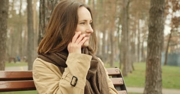 Retrato de la mujer hablando por teléfono sentado en el banco en el parque de otoño con bufanda marrón disfrutando de la conversación . — Vídeos de Stock