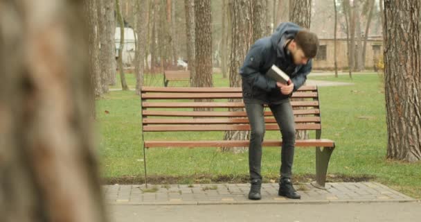 Όμορφος γενειοφόρος κάθεται στο παγκάκι κρατώντας ένα βιβλίο, ανοίγει ένα βιβλίο γυρίζει σελίδες και αρχίζει να διαβάζει. Ανάγνωση στο πάρκο — Αρχείο Βίντεο