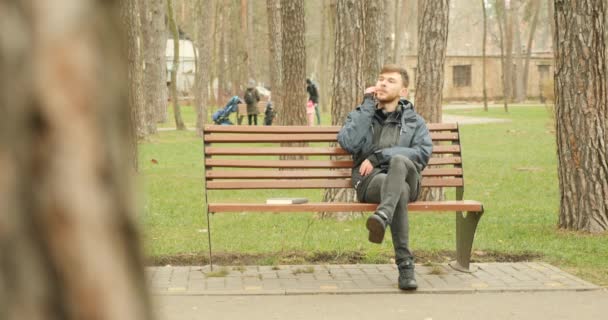 秋の公園のベンチに座って会話を楽しみながら笑顔で電話で話し始める若い髭の男. — ストック動画