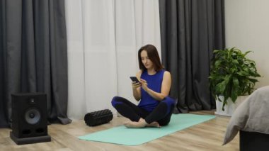 Woman with smartphone meditating. Young female in sports clothing pantone color relaxing by practicing yoga. Woman sitting on yoga mat and posing lotus finger gesture at home. Slow motion