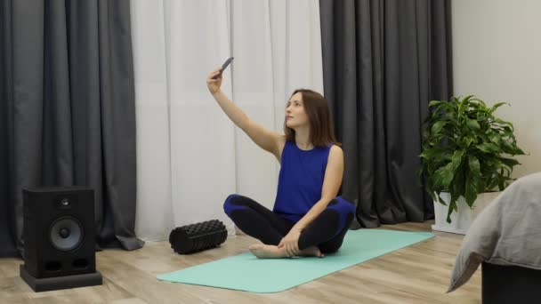 Frau macht Selfie auf Yogamatte zu Hause. junge kaukasische Frau, die in Zen-Position auf dem Boden sitzt und meditiert. Unbekümmertes erwachsenes Mädchen beim Telefonieren vor dem Yoga-Üben — Stockvideo