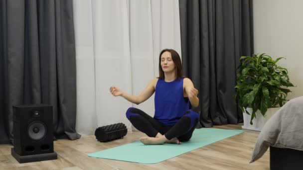 Vrouw mediterend met gesloten ogen en handen in namaste op mat thuis. Ontspannen vrouw in sportkleding pantalon kleur ontspannen in lotus positie zittend op yoga mat. Spiritualiteit levensstijl concept — Stockvideo