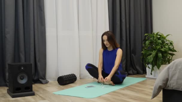 La mujer se pone auriculares y se prepara para practicar yoga. Mujer joven caucásica en pantone ropa deportiva azul sentada en una esterilla de yoga y poniéndose auriculares antes de meditar. Concepto de atención plena — Vídeos de Stock