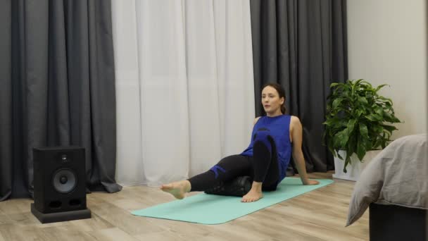 Frau führt myofasziale Freisetzung mit Fitnessroller durch. junge kaukasische Brünette tut erholsame und entspannende Massage auf Yogamatte im hellen Wohnzimmer. fitte Frau macht Pilates Reha-Übungen — Stockvideo