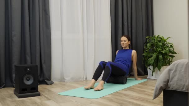 Güzel bir kadın yoga minderinde fasya rulosu kullanarak kalça kaslarına masaj yapar. Mavi bluzlu ve tozluklu genç bir kadın spor yaptıktan sonra rehabilitasyon ve rehabilitasyon egzersizleri yapıyor. Ev fitness konsepti — Stok video