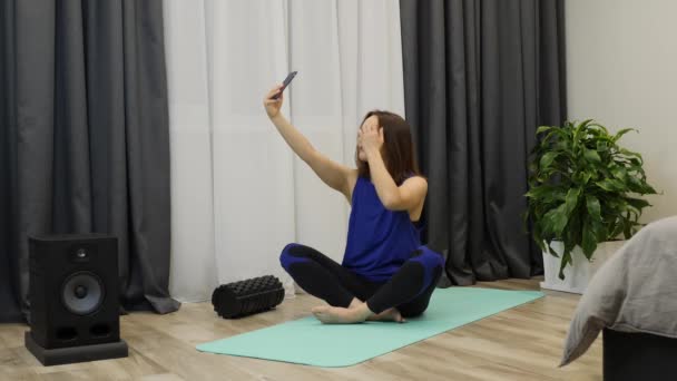 Frau macht Selfie auf Yogamatte zu Hause. junge kaukasische Frau, die in Zen-Position auf dem Boden sitzt und meditiert. Unbekümmertes erwachsenes Mädchen, das vor dem Yoga am Telefon Fotos macht. Zeitlupe — Stockvideo