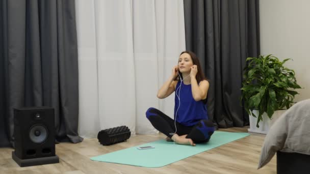 Female in earphones starts meditation sitting on yoga mat at home. Relaxed young carefree woman meditating with earphones in ears. Woman listening to relaxing music during yoga lessons. Slow motion — Stockvideo