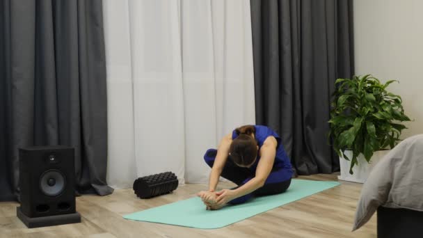 Vrouw strekt lichaamsspieren uit. Ontspannen geconcentreerde vrouw doet stretching oefeningen op yoga mat. Brunette in pantalon blauwe kleren die heup uitrekt. Sportief meisje doet fitness training. Langzame beweging — Stockvideo