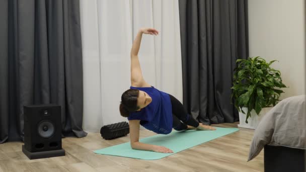 Mulher a fazer prancha lateral. Mulher concentrada fazendo alongamento exercício de ioga no tapete em casa. Ajuste morena fazendo exercícios de treino abdominal. Menina em sportswear exercício no tapete de ioga. Movimento lento — Vídeo de Stock