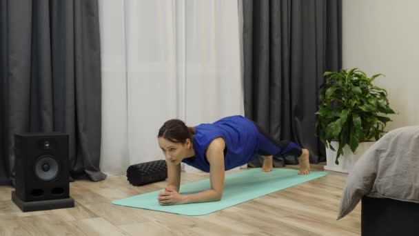 Девушка позирует на коврике для йоги. Молодая спортсменка делает доску в рамках ежедневных упражнений. Женщина в голубой одежде в пантоне стоит на доске. Медленное движение — стоковое видео