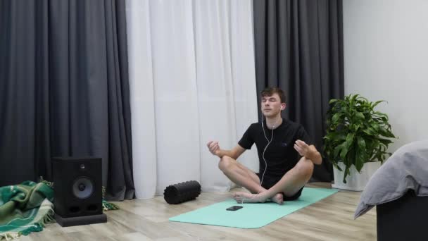 Człowiek w słuchawkach medytuje. Młody mężczyzna w sportowej odzieży ćwiczący jogę. kaukaski chłopiec siedzi na macie do jogi i pozowanie lotosu palec gest w domu — Wideo stockowe