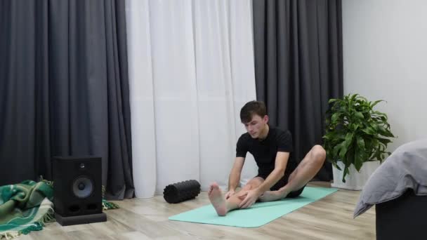 Masculino estira los músculos de la cadera, músculos de las piernas en estera de yoga. Hombre gimnasta en ropa negra se está estirando en casa. Niño caucásico haciendo ejercicios de fitness y yoga. Concepto de salud y estilo de vida — Vídeos de Stock