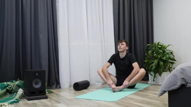 Manlig idrottsman sträcker sig hemma. Ung kaukasisk idrottsman i svarta kläder gör yogaövningar på yogamattan. Snygg pojke som tränar fitness och aerob träning i vardagsrummet. Män som tränar hemma — Stockvideo