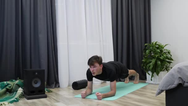 El atleta masculino posa sobre una alfombra. Joven hombre en forma haciendo tablón como parte de sus ejercicios diarios. Entrenador de fitness en ropa negra está en posición de tablón. Concepto de entrenamientos — Vídeos de Stock