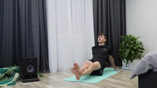 Człowiek robi ćwiczenia w salonie. Chłopak masuje mięśnie biodra używając rolki do jogi. Instruktor fitness robi ćwiczenia rehabilitacyjne po ciężkim treningu w domu. mężczyzna robi masaż z fascia roll — Wideo stockowe