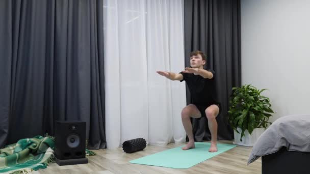 Entrenador de fitness hace sentadillas en interiores. Hombre atlético confiado haciendo ejercicios de sentadilla en casa. Hombre en forma joven está en cuclillas en la esterilla de yoga. Un hombre haciendo ejercicios de sentadillas. Boy hace entrenamiento aeróbico — Vídeos de Stock